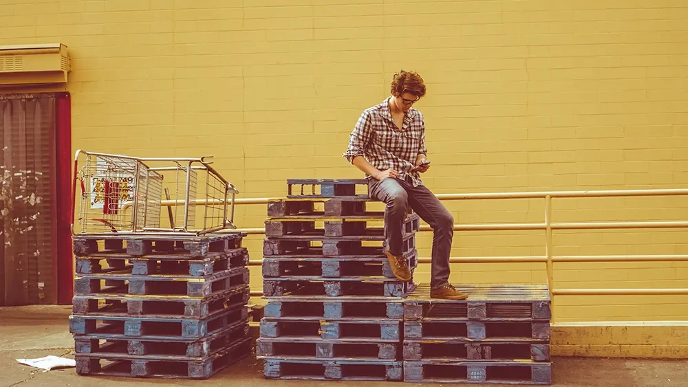 Man sitting on pallets, reading phone, wasting time.